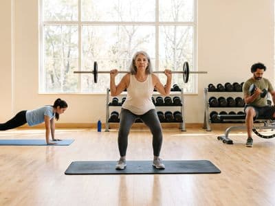 A bright gym floor seen wide, with three ordinary people working separately on different things — a bar, a bench, a mat — plenty of light and space