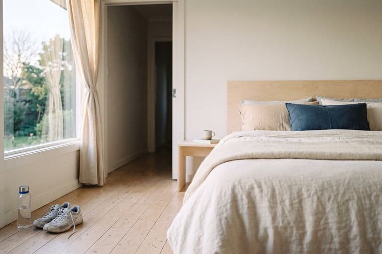 A quiet bedroom in early morning light with curtains half open and a made bed, running shoes and a water bottle by the door. Nobody in the frame