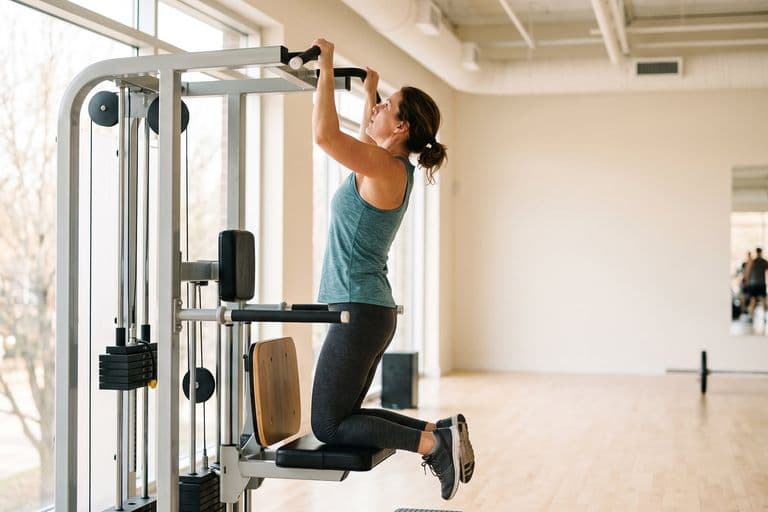 An ordinary woman in her forties of average build mid-set on an assisted pull-up machine, focused and slightly out of breath, looking up at the bar