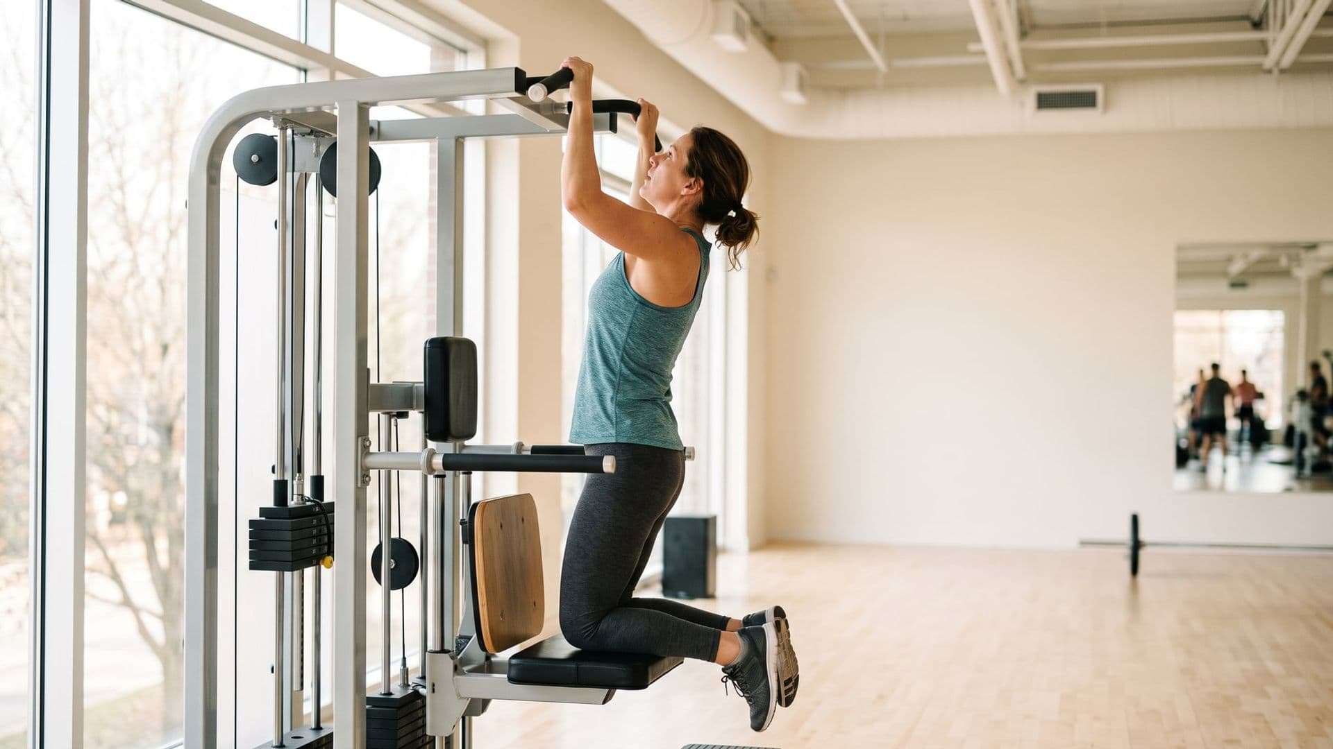 An ordinary woman in her forties of average build mid-set on an assisted pull-up machine, focused and slightly out of breath, looking up at the bar